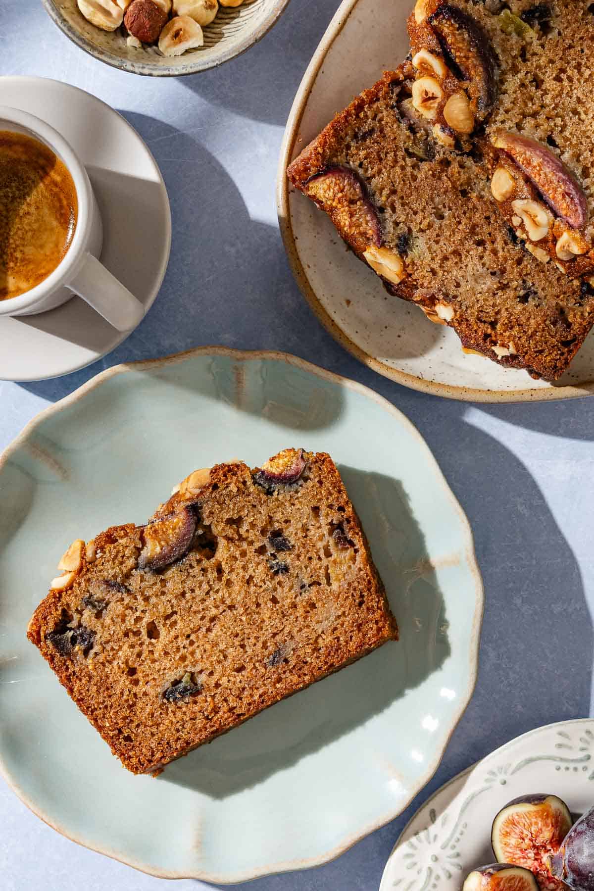 An overhead photo of a slice of whole wheat fig bread on a plate. Next to this is a cup of coffee, bowls of hazelnuts and figs, and a serving plate with more slices of the bread.