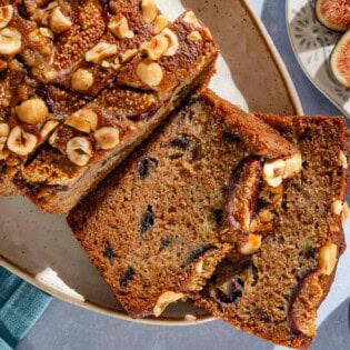 An overhead photo of a loaf of partially sliced whole wheat fig bread on a serving plate. Next to this are bowls of hazelnuts and figs.