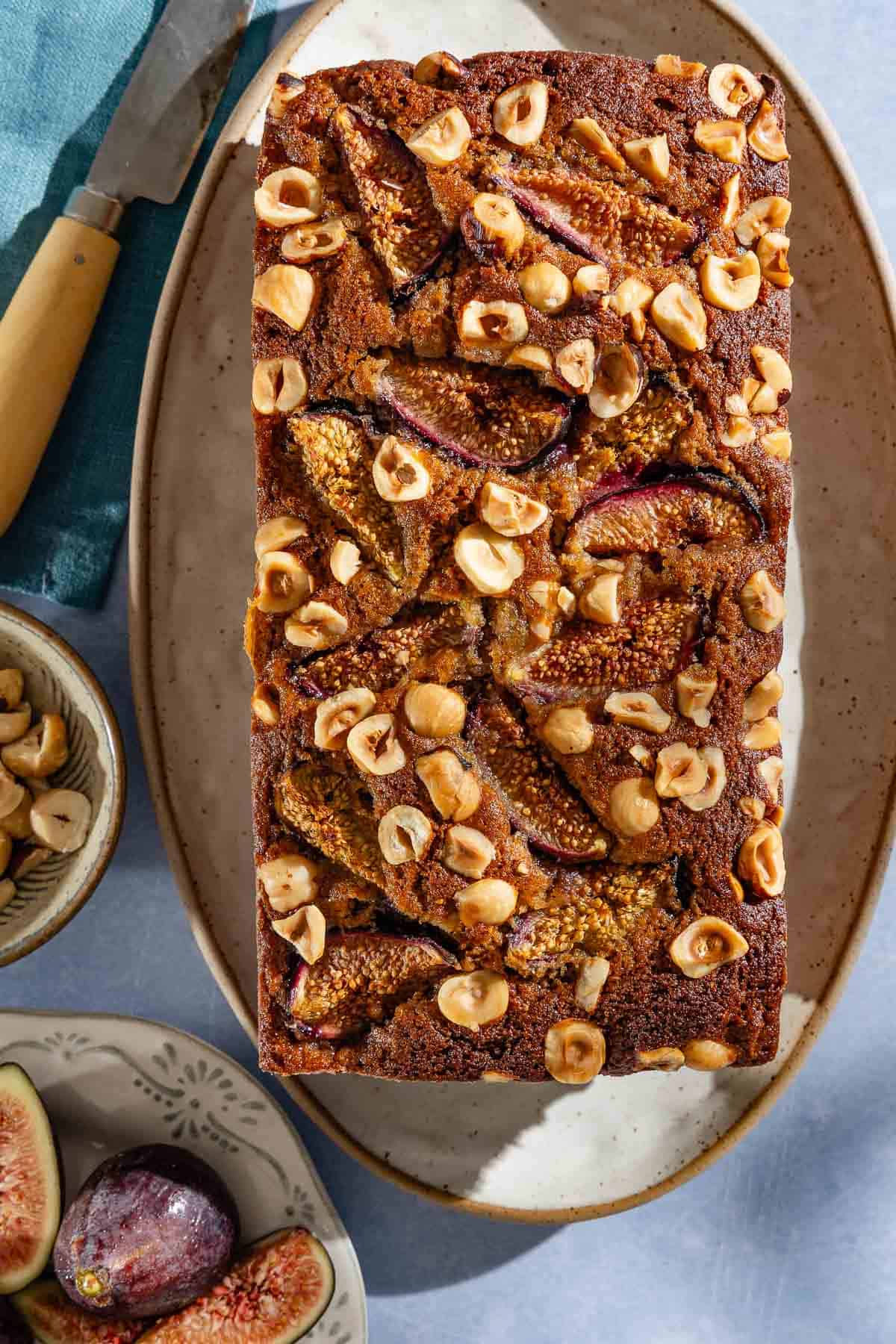 An overhead photo of a loaf of whole wheat fig bread on a serving plate. Next to this is a knife and bowls of hazelnuts and figs.