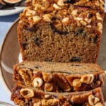 A close up of a loaf of partially sliced whole wheat fig bread on a serving plate. Next to this are bowls of hazelnuts and figs.