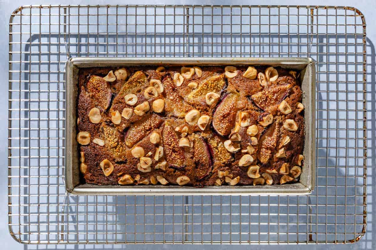 An overhead photo of a loaf of whole wheat fig bread in a baking pan cooling on a wire rack.