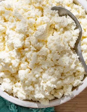 An overhead close up photo of homemade ricotta in a bowl with a spoon next to a kitchen towel.