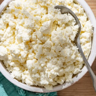 An overhead close up photo of homemade ricotta in a bowl with a spoon next to a kitchen towel.