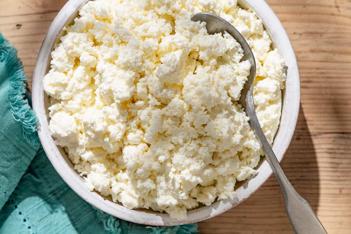 An overhead close up photo of homemade ricotta in a bowl with a spoon next to a kitchen towel.