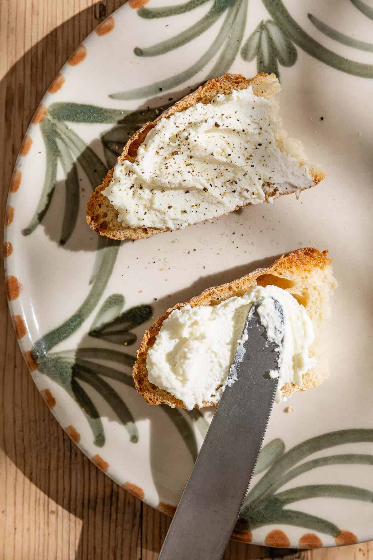 An overhead photo of homemade ricotta being spread on 2 pieces of bread with a knife.