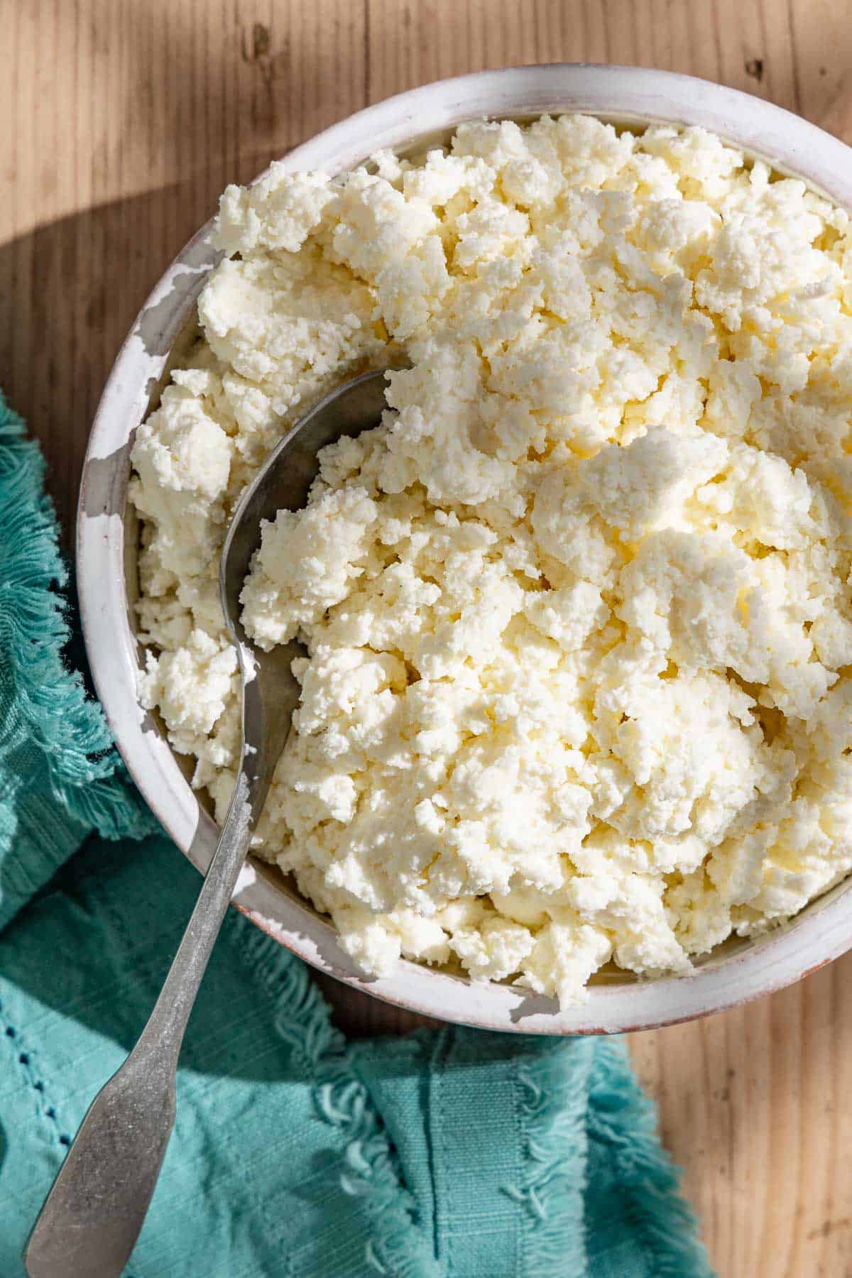 An overhead photo of homemade ricotta in a bowl with a spoon next to a kitchen towel.