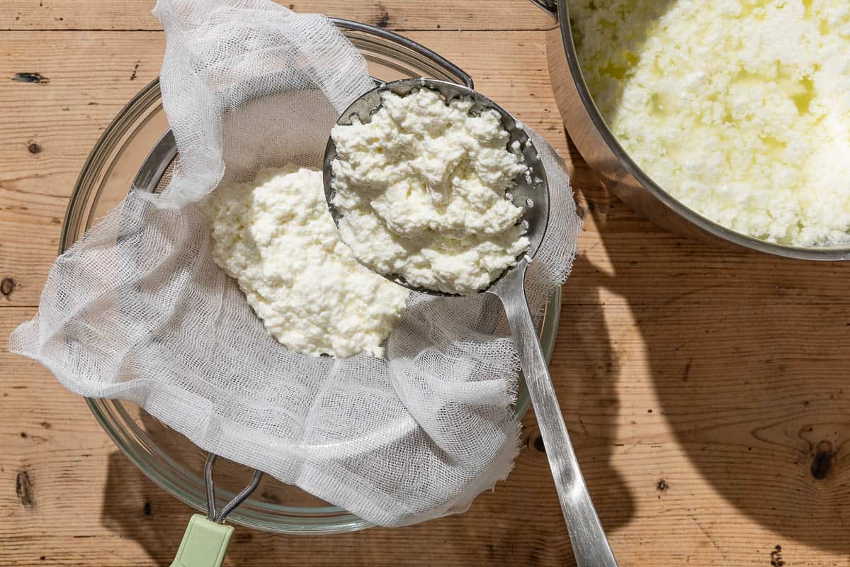 Homemade ricotta being ladled from a large pot into in a cheesecloth-lined mesh strainer sitting over a bowl.