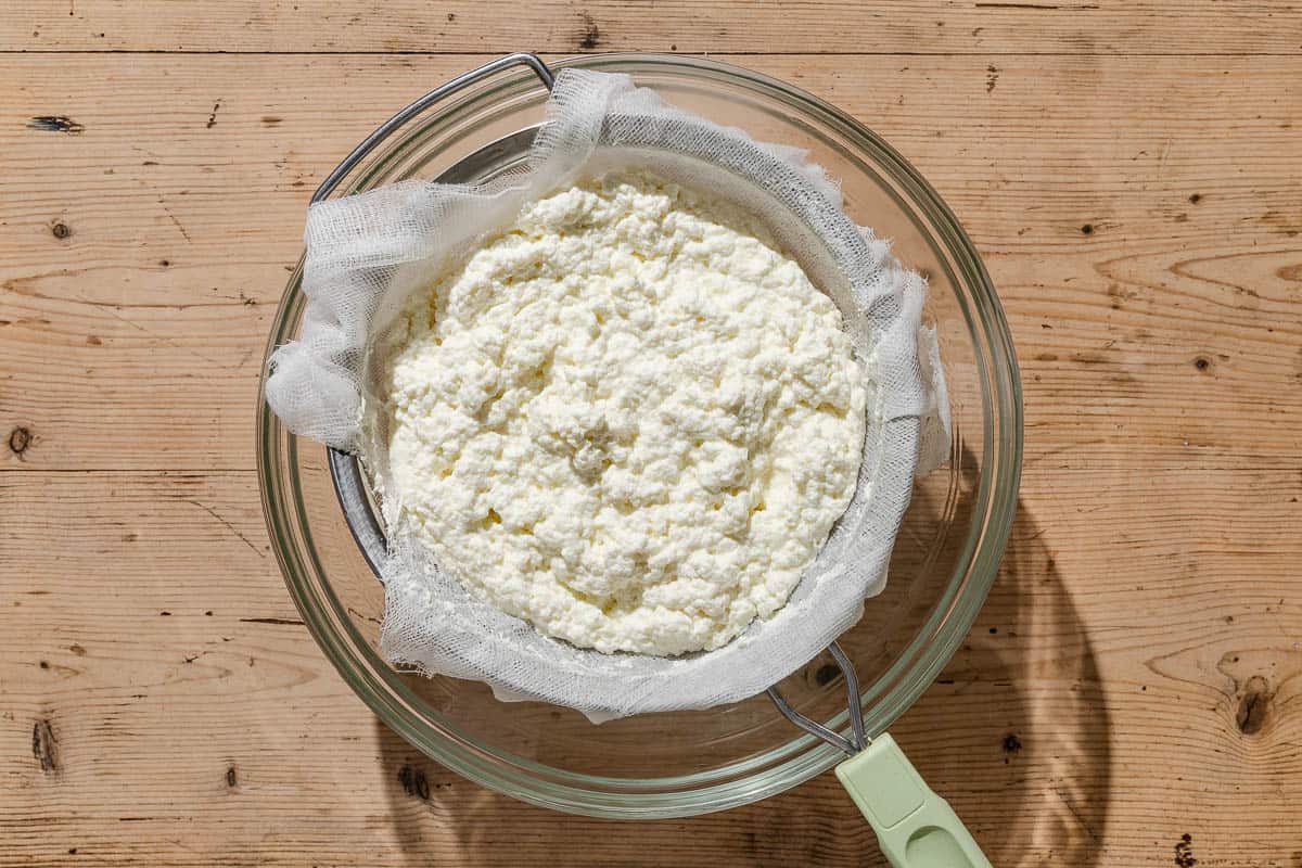 Homemade ricotta being strained in a cheesecloth-lined mesh strainer sitting over a bowl.