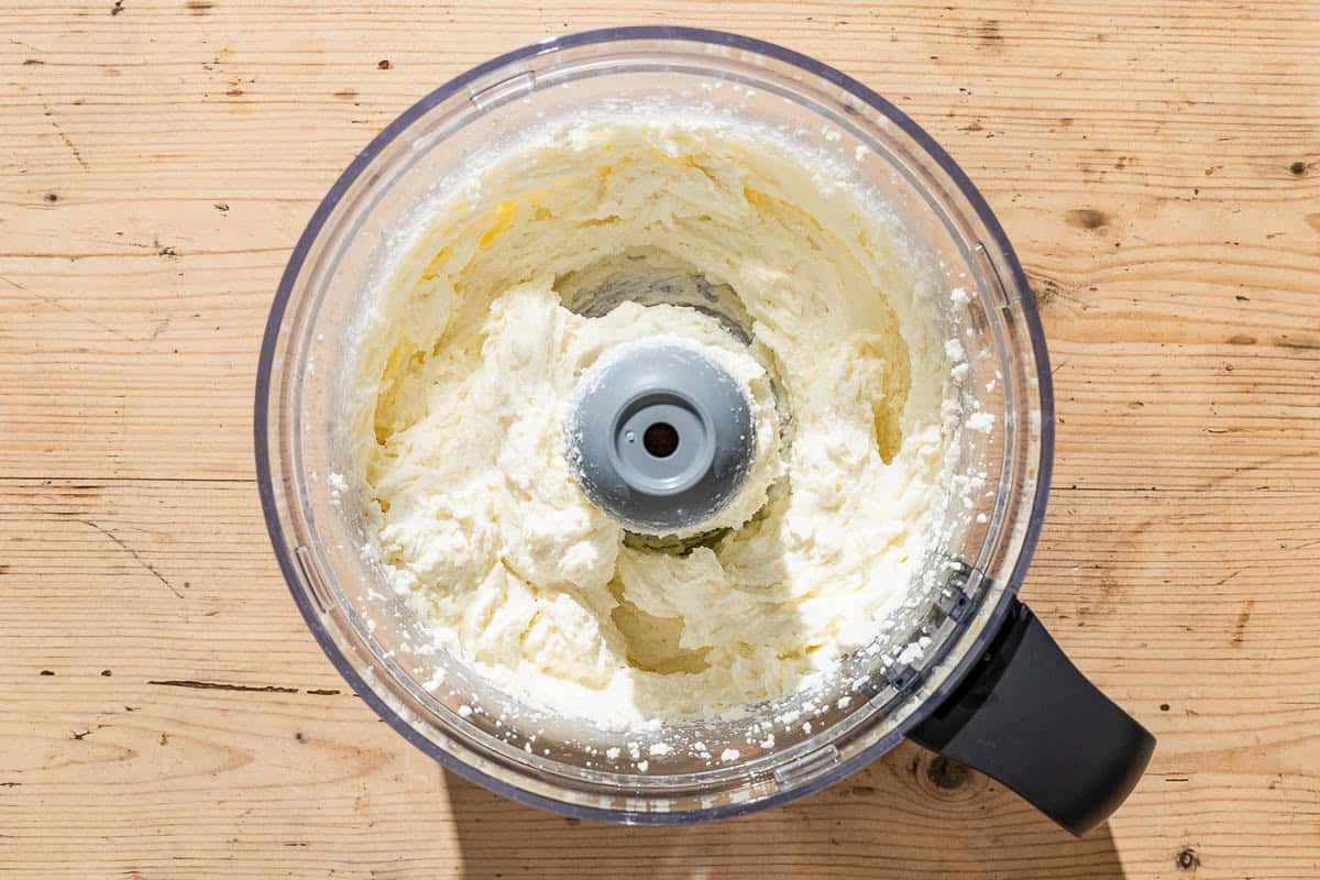 An overhead photo of homemade ricotta in the bowl of a food processor fitted with a blade.