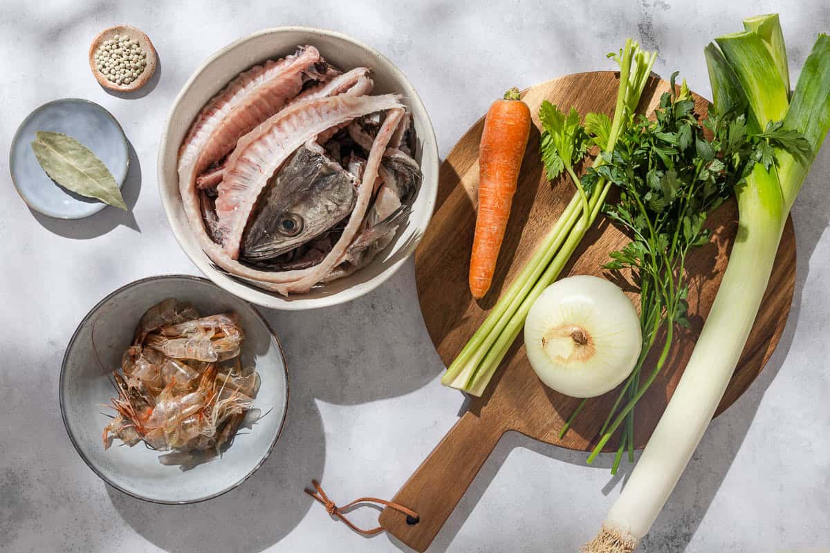 An overhead photo of the ingredients for the seafood stock also called seafood broth including a bowl of fish bones, raw crustacean shells, an onion, a leek, a carrot, celery, parsley, white peppercorns and a bay leaf.