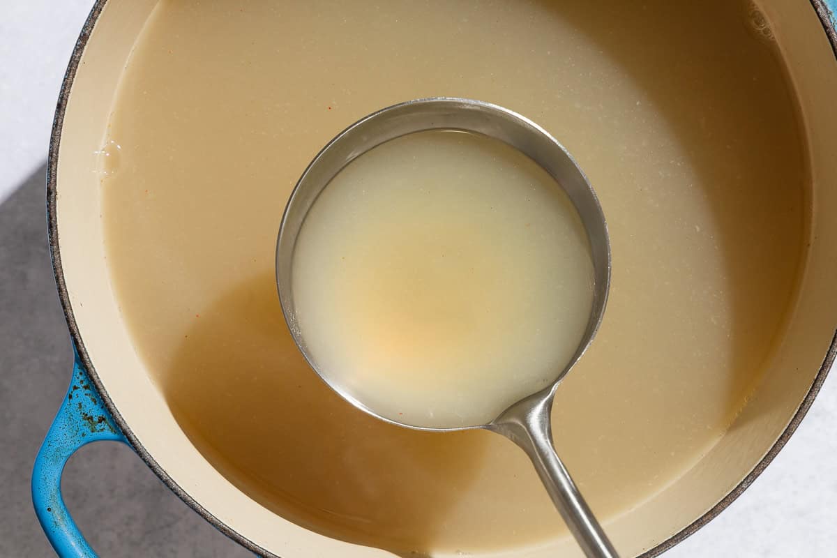 An overhead photo of seafood stock in a ladle being held over a pot of the stock.