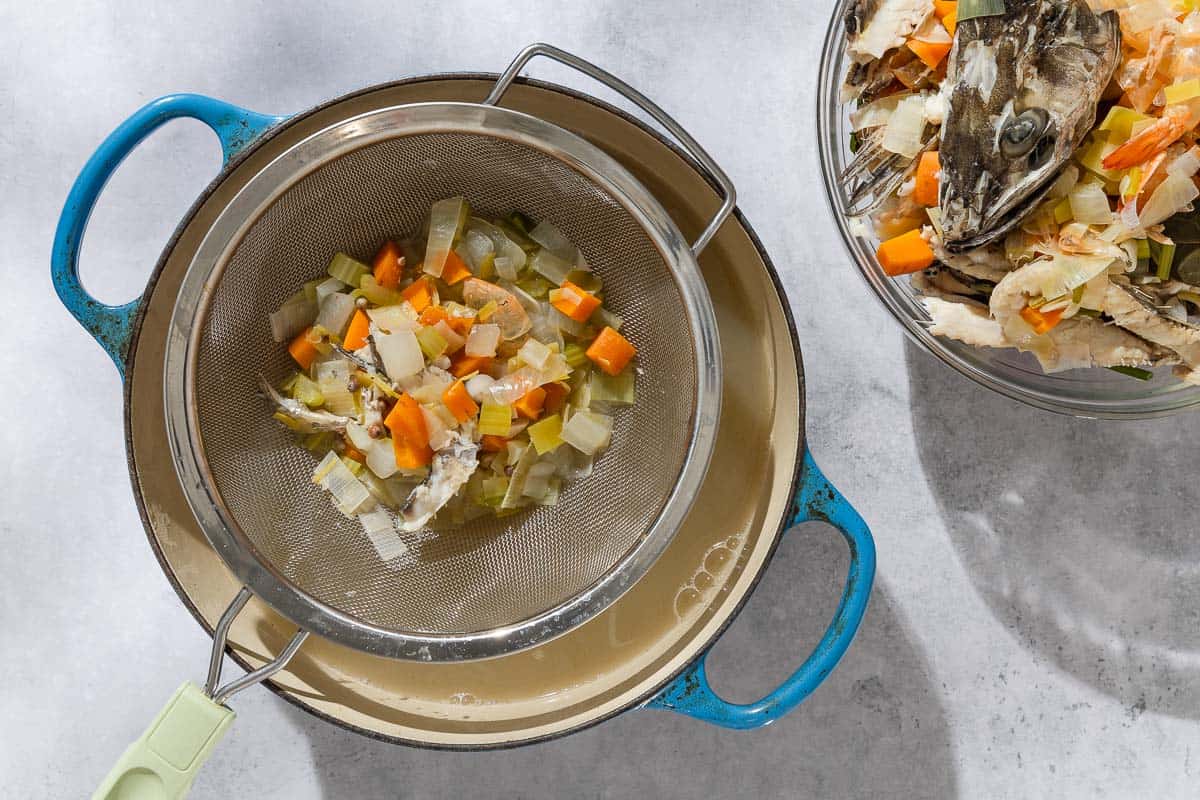 An overhead phot of the seafood stock being strained with a mesh strainer held over a pot. Next to this is more of the strained ingredients in a bowl.