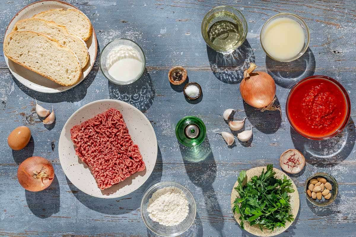 Ingredients for Spanish meatballs including slices of bread, milk, ground beef, onion, garlic, egg, parsley, salt, black pepper, flour, olive oil, crushed tomatoes, almonds, saffron, white wine and chicken stock.