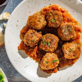 An overhead photo of a serving of Spanish meatballs and sauce on a plate. Next to this is a slice of bread and a glass of red wine.