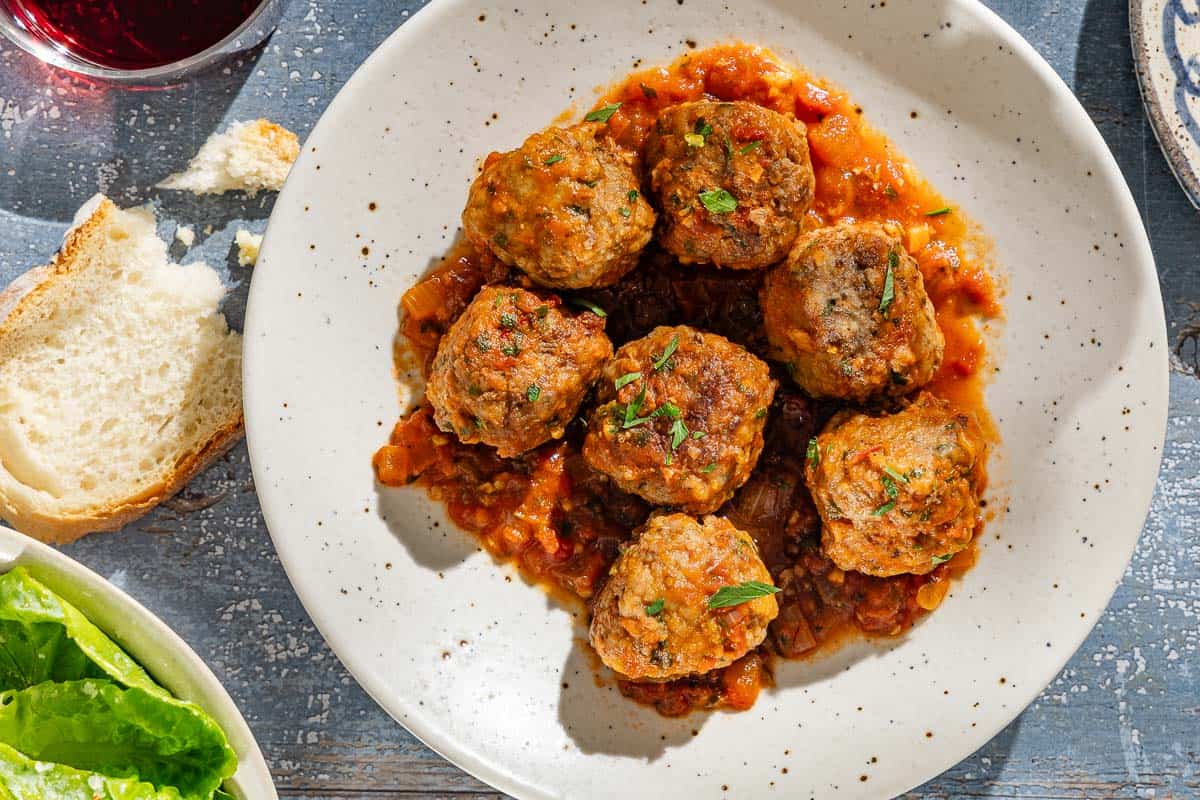 An overhead photo of a serving of Spanish meatballs and sauce on a plate. Next to this is a slice of bread and a glass of red wine.