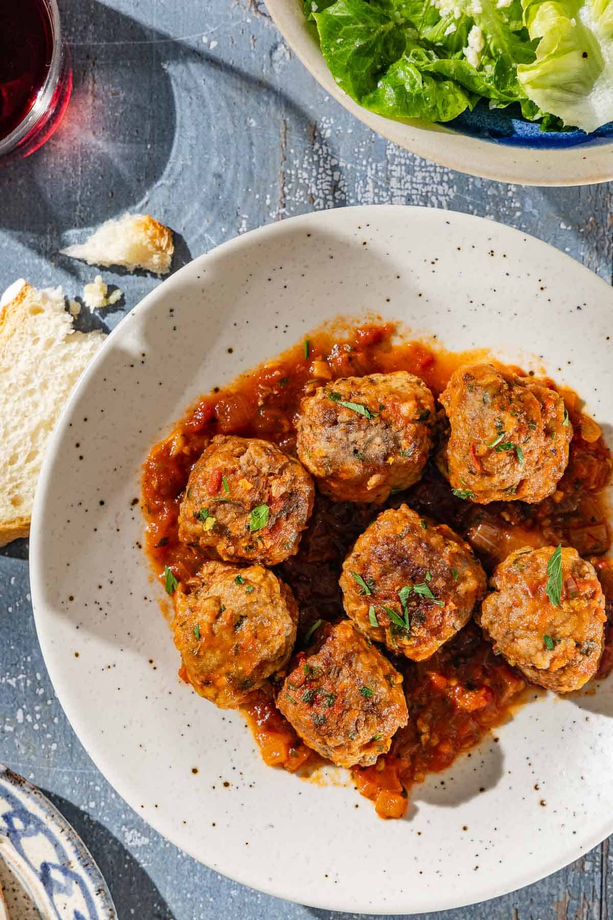 An overhead photo of a serving of Albóndigas (Spanish meatballs) and sauce on a plate. Next to this a glass of red wine, a bowl of salad and a another slice bread.