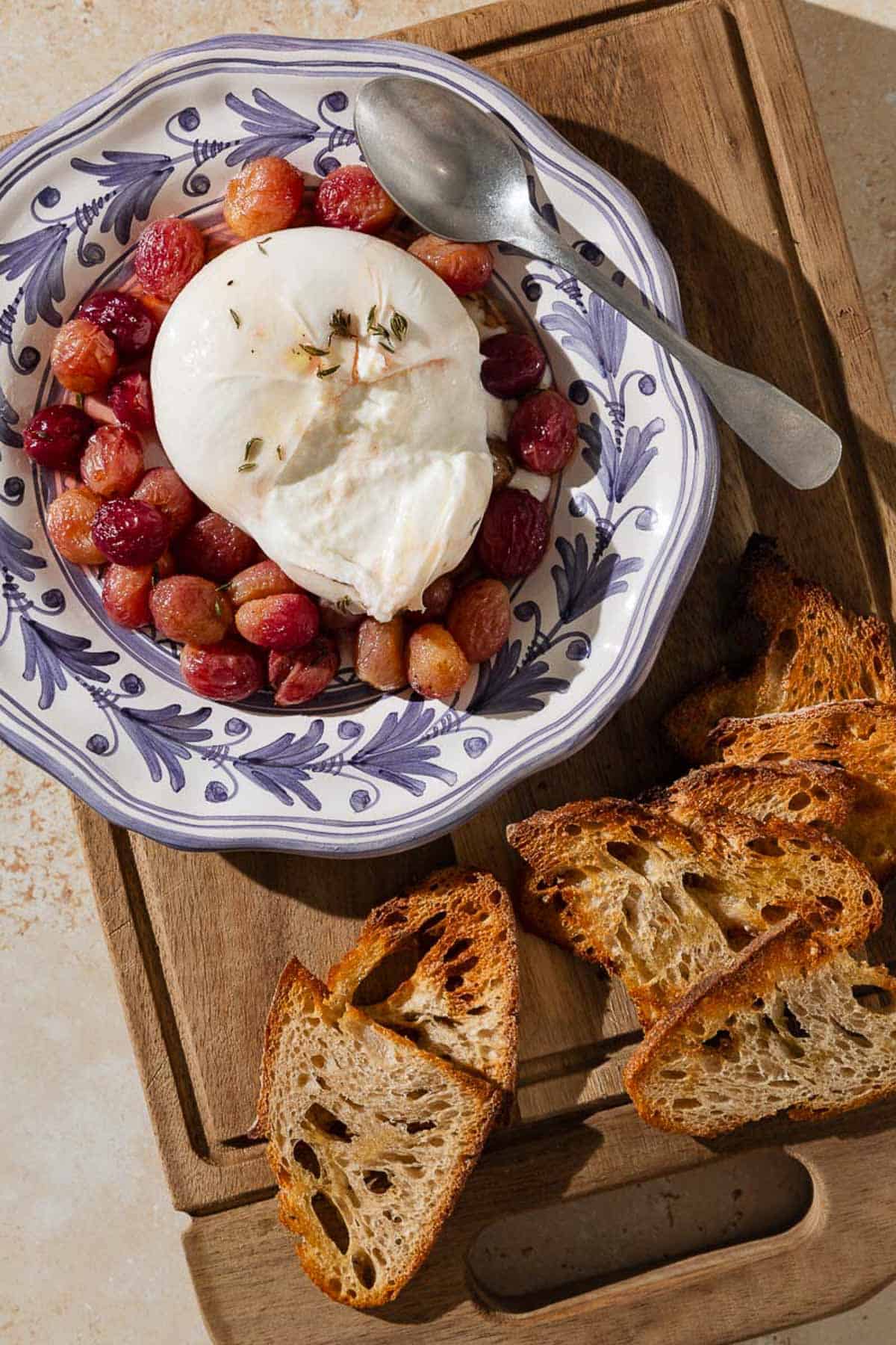 An overhead photo of roasted grapes topped with burrata, sherry and thyme in a bowl with a spoon next to slices of toasted bread on a wooden tray.