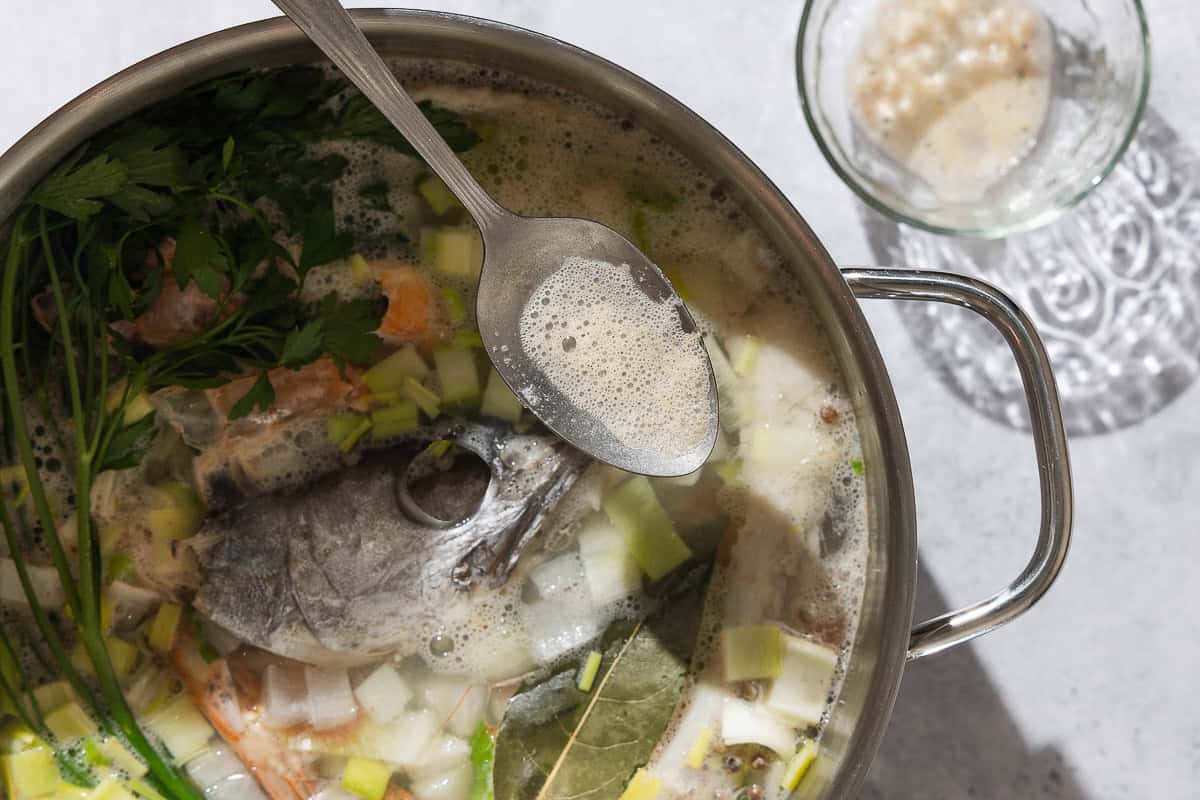 A close up photo of a bit of foam from the seafood stock on a spoon being held over the large pot of the stock. Next to this is a small bowl with more of the foam.