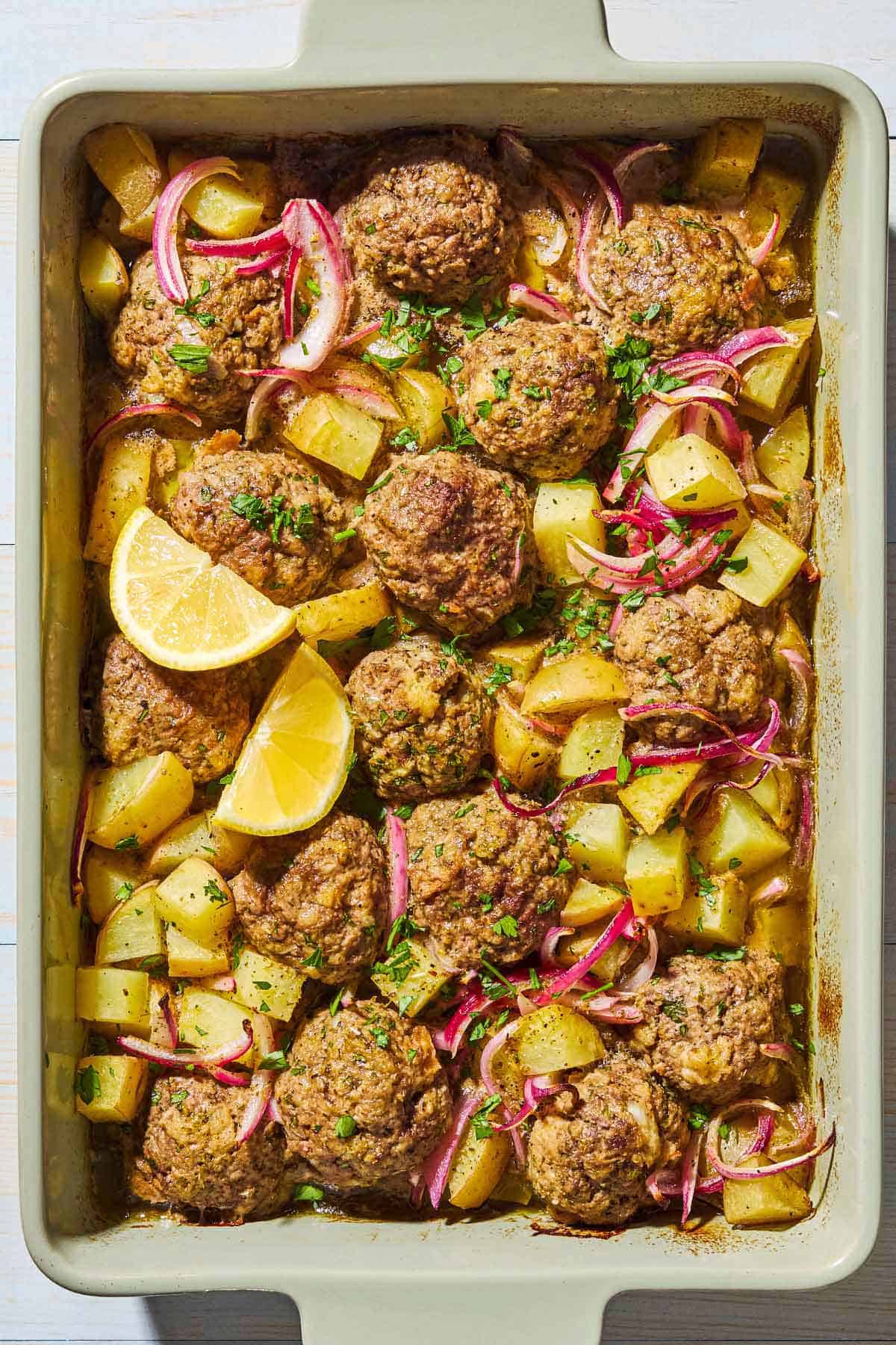 An overhead photo of baked greek meatballs and potatoes in a baking dish with lemon wedges.