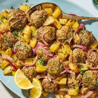 Baked greek meatballs and potatoes on a serving plate with a serving spoon and lemon wedges. Next to this is a small bowl of chopped parsley.