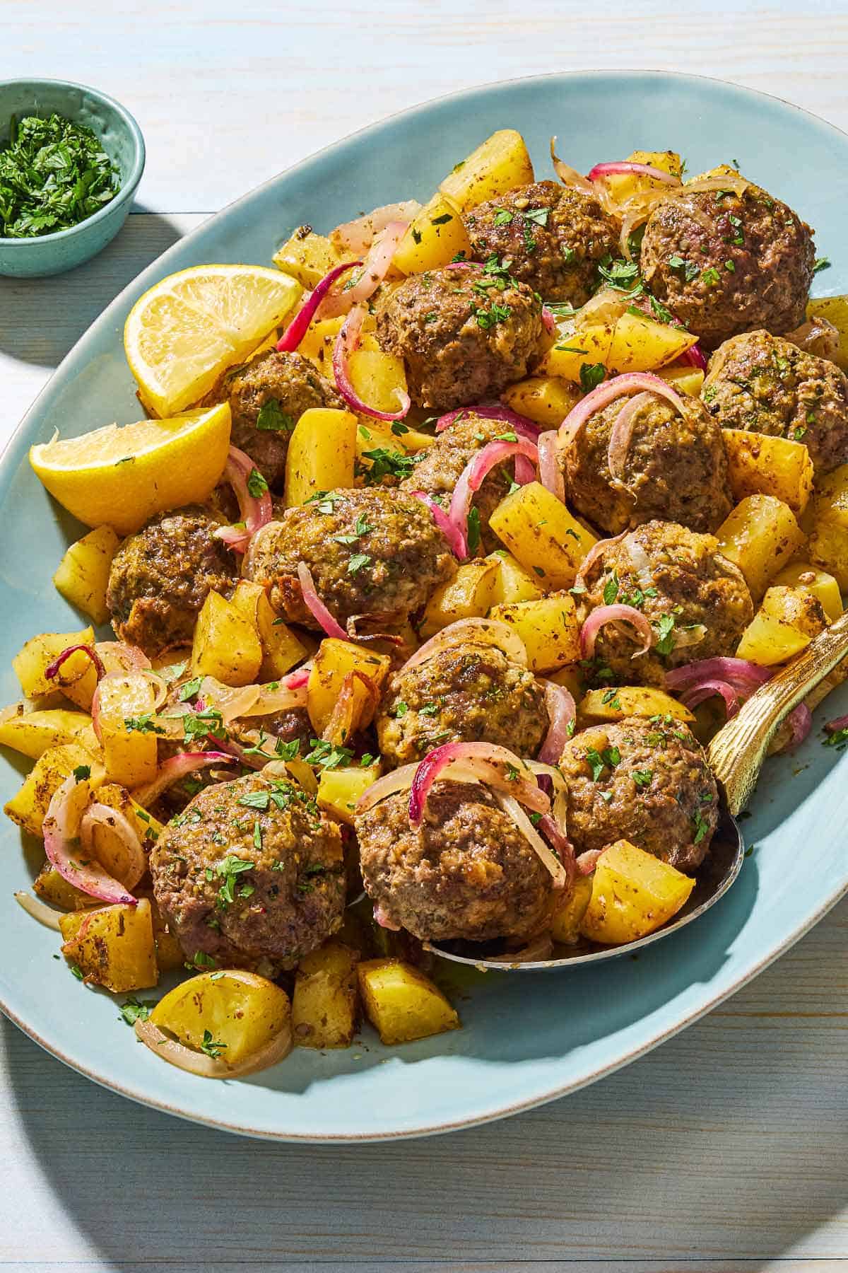 Baked greek meatballs and potatoes on a serving plate with a serving spoon and lemon wedges. Next to this is a small bowl of chopped parsley.