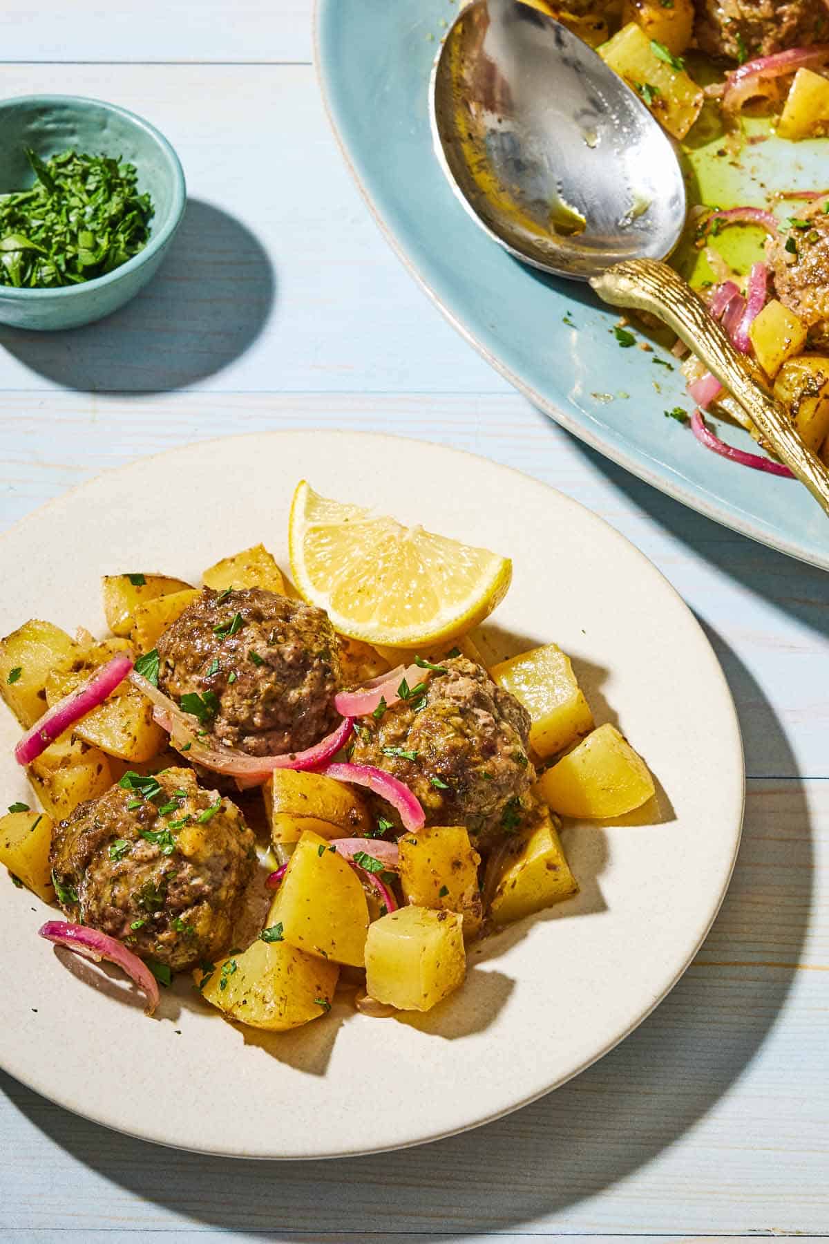 Baked greek meatballs and potatoes on a plate. Next to this is a small bowl of chopped parsely and the rest of the baked meatballs an potatoes on a serving platter with a spoon.