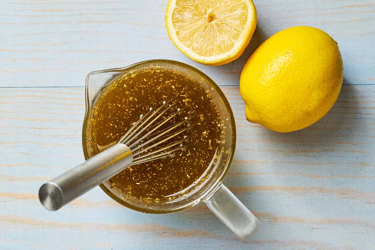 Ladolemono greek salad dressing in a measuring cup with a whisk. Next to this is a whole lemon and a lemon half.