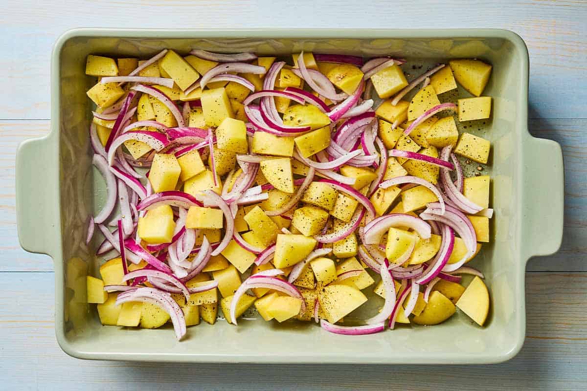 An overhead photo of uncooked chopped potatoes and red onions seasoned with salt and pepper in a baking dish.