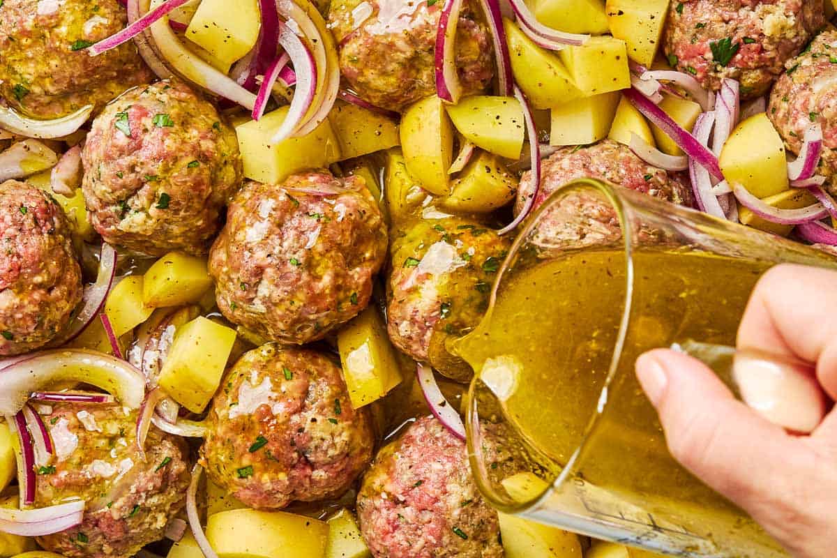 An overhead photo of the ladolemono sauce being poured on the uncooked greek meatballs and potatoes.