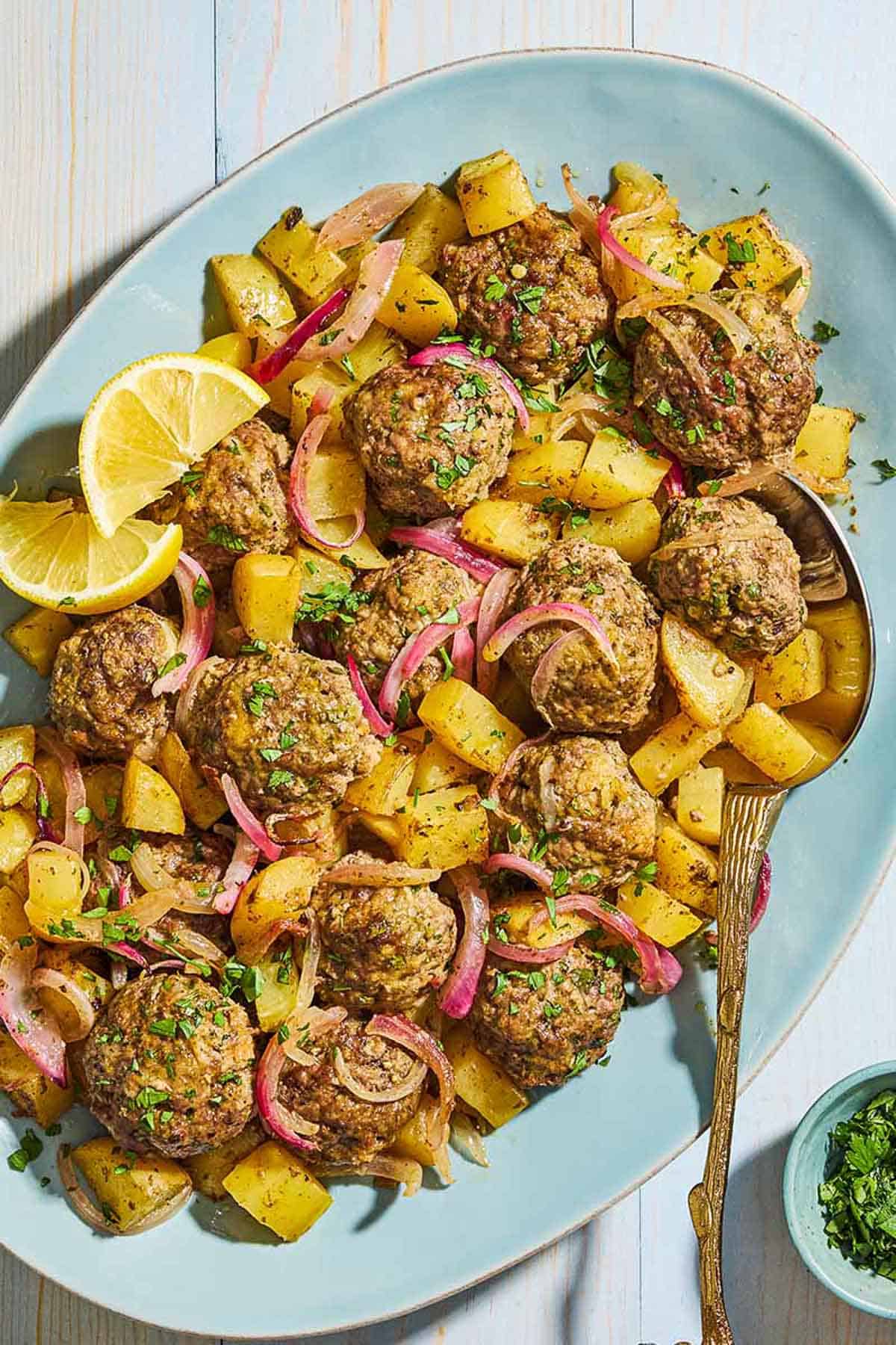 An overhead photo of baked greek meatballs and potatoes on a serving plate with a serving spoon and lemon wedges. Next to this is a small bowl of chopped parsley.