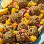 A close up of baked greek meatballs and potatoes on a serving plate with a serving spoon and lemon wedges.