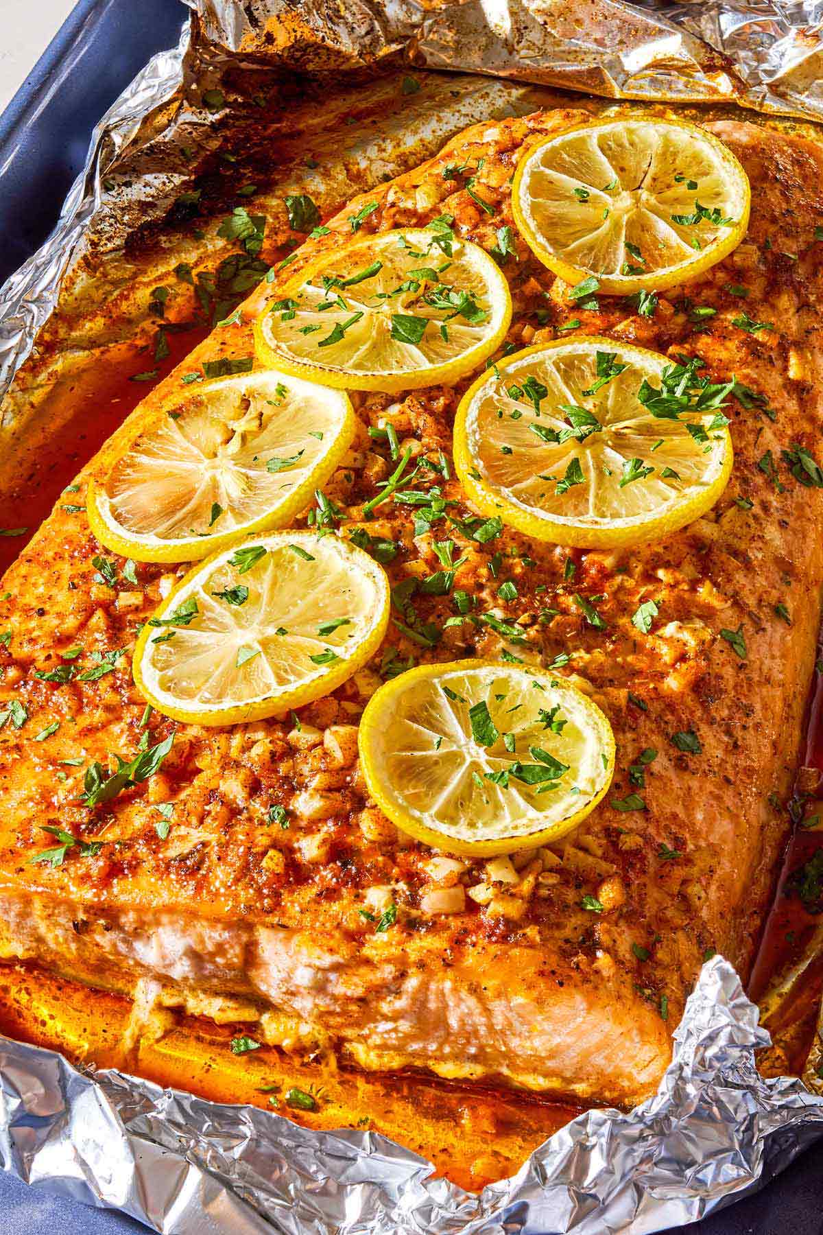 A close up photo of a lemon garlic salmon fillet topped with lemon wheels and parsley on an aluminum foil-lined sheet pan.