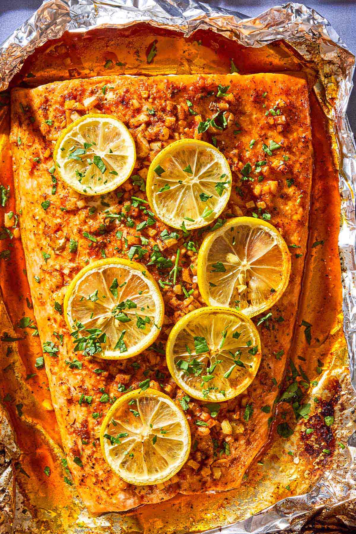 An overhead close up photo of a lemon garlic salmon fillet topped with lemon wheels and parsley on an aluminum foil-lined sheet pan.