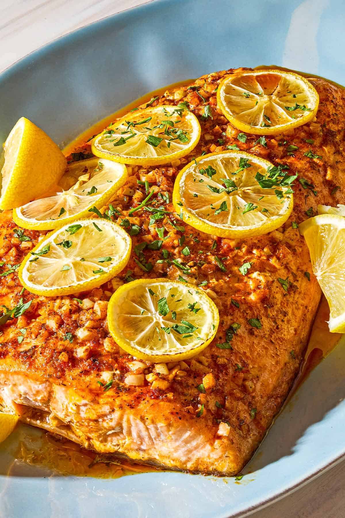 A close up photo of a lemon garlic salmon fillet topped with lemon wheels and parsley on a serving platter with lemon wedges.