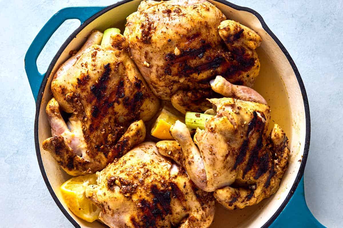 An overhead photo of 4 browned cornish hens in a large skillet with lemon wedges.