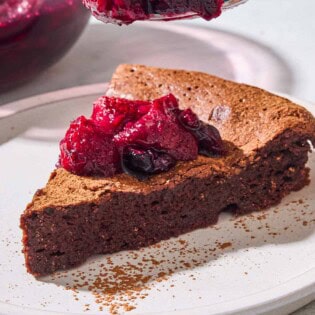 A close up of a slice of flourless chocolate cake topped with berry compote on a plate with a spoonful of the compote being held above it. Behind this is a bowl of the rest of the compote.