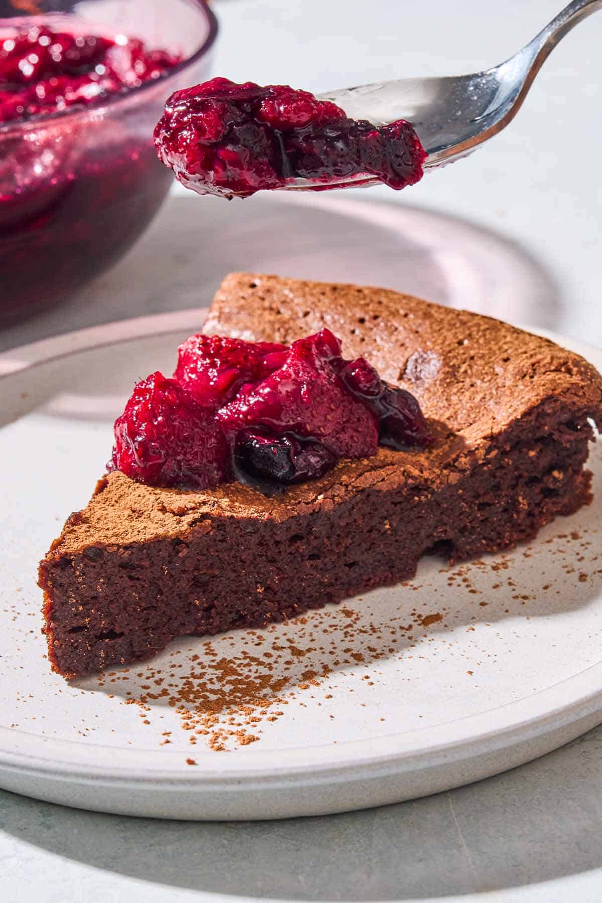 A close up of a slice of flourless chocolate cake topped with berry compote on a plate with a spoonful of the compote being held above it. Behind this is a bowl of the rest of the compote.