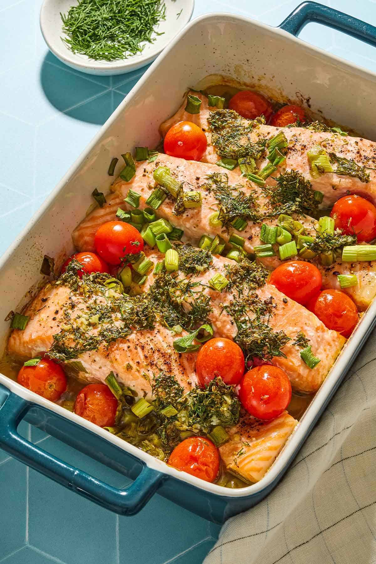 4 Greek salmon fillets with cherry tomatoes in a baking dish. Next to this is a small bowl of dill.