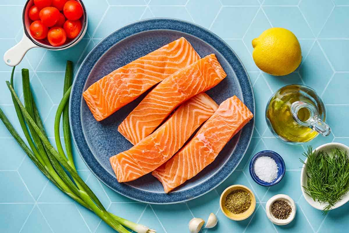 Ingredients for Greek salmon including salmon fillets, lemon, garlic, oregano, fill, salt, black pepper, green onions and cherry tomatoes.