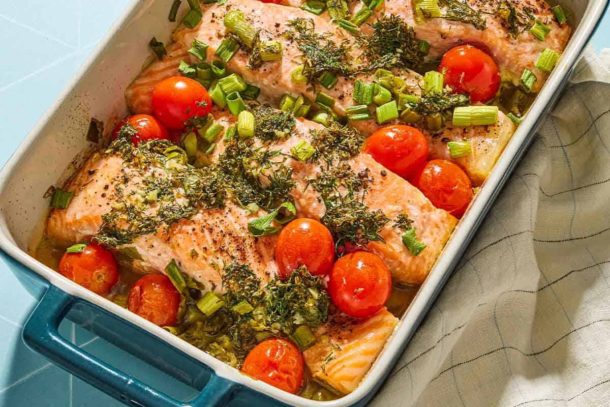 A close up of 4 Greek salmon fillets with cherry tomatoes in a baking dish. Next to this is a cloth napkin.