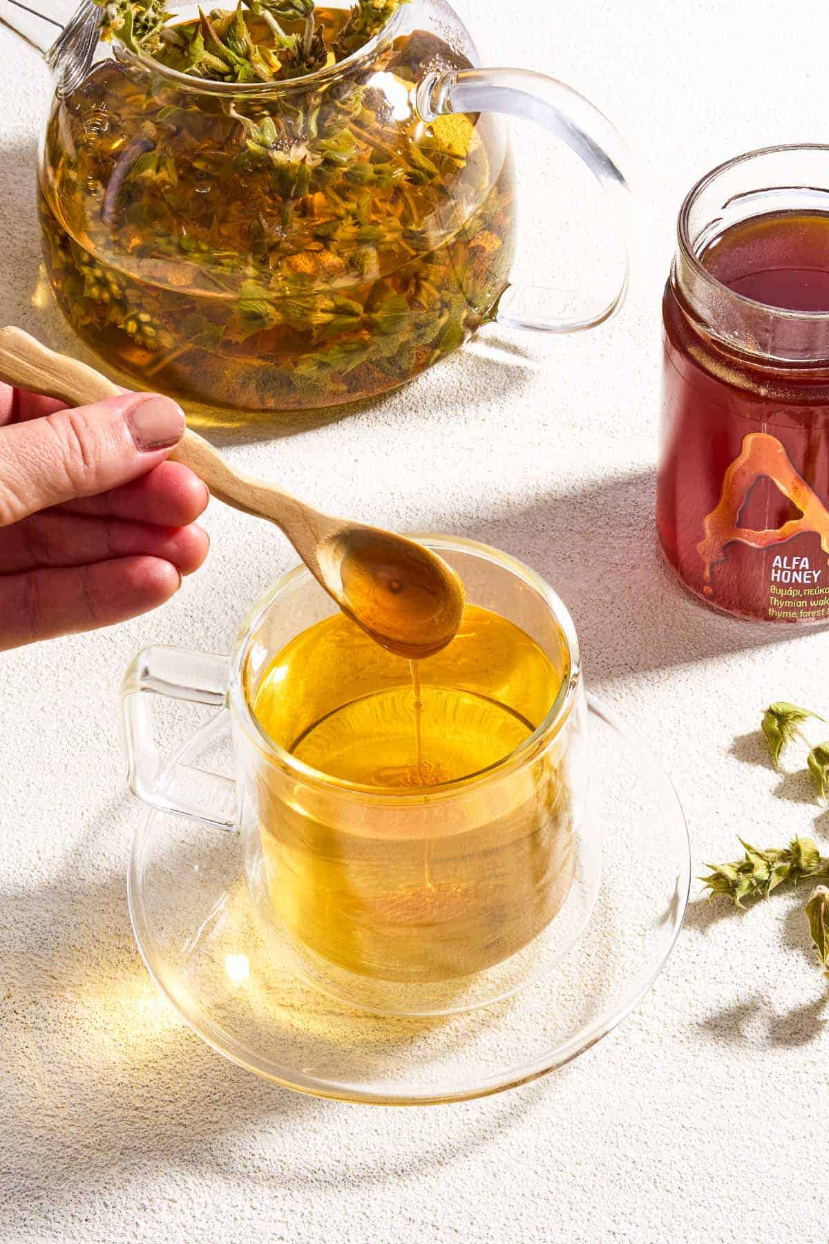 A close up of honey being spooned into a cup of Greek mountain tea on a saucer. Next to this is the kettle with the rest of the tea, and a jar of honey.