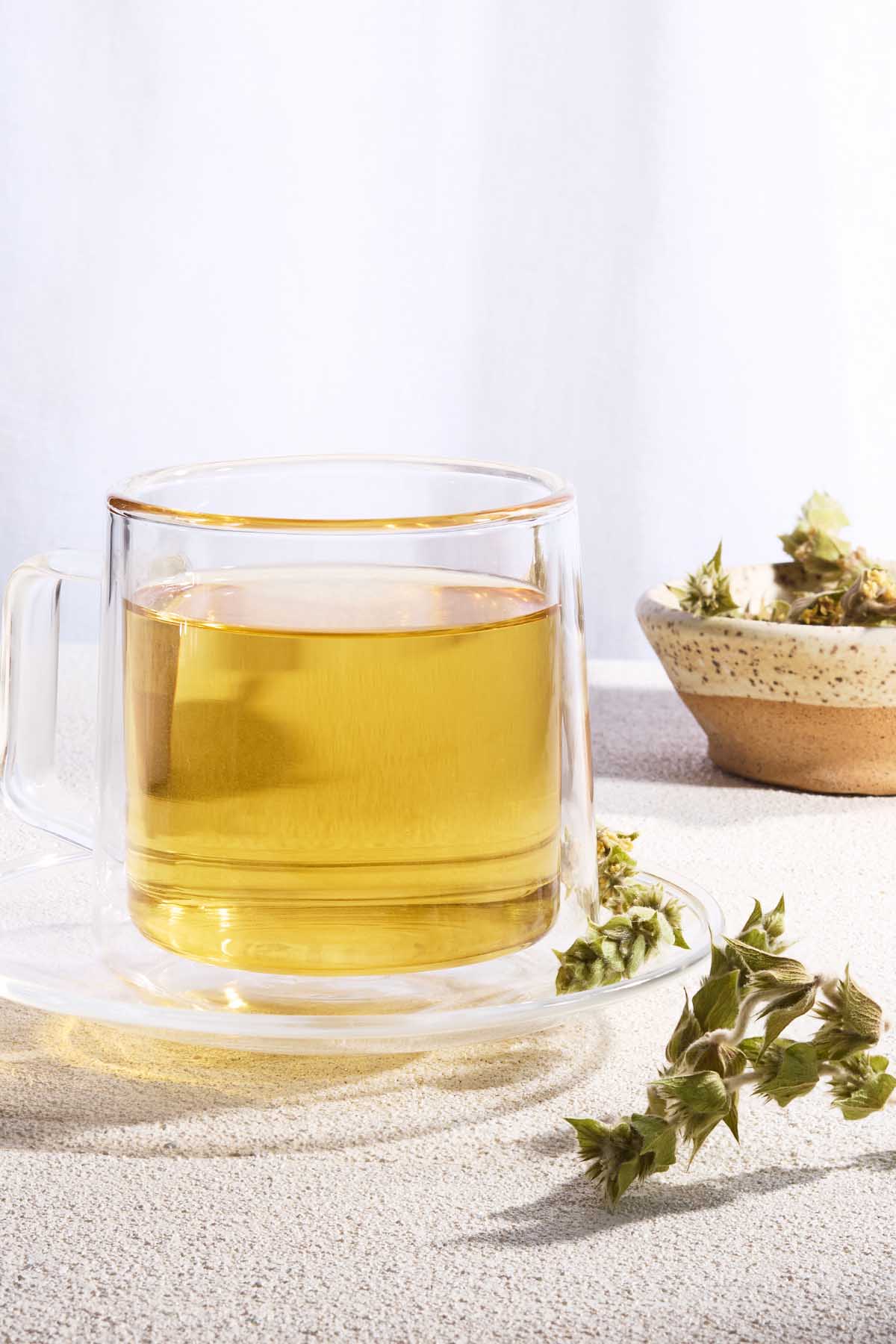 A close up of a cup of brewed Greek mountain tea on a saucer. Next to this is a bowl of the tea leaves and a sprig of the tea.