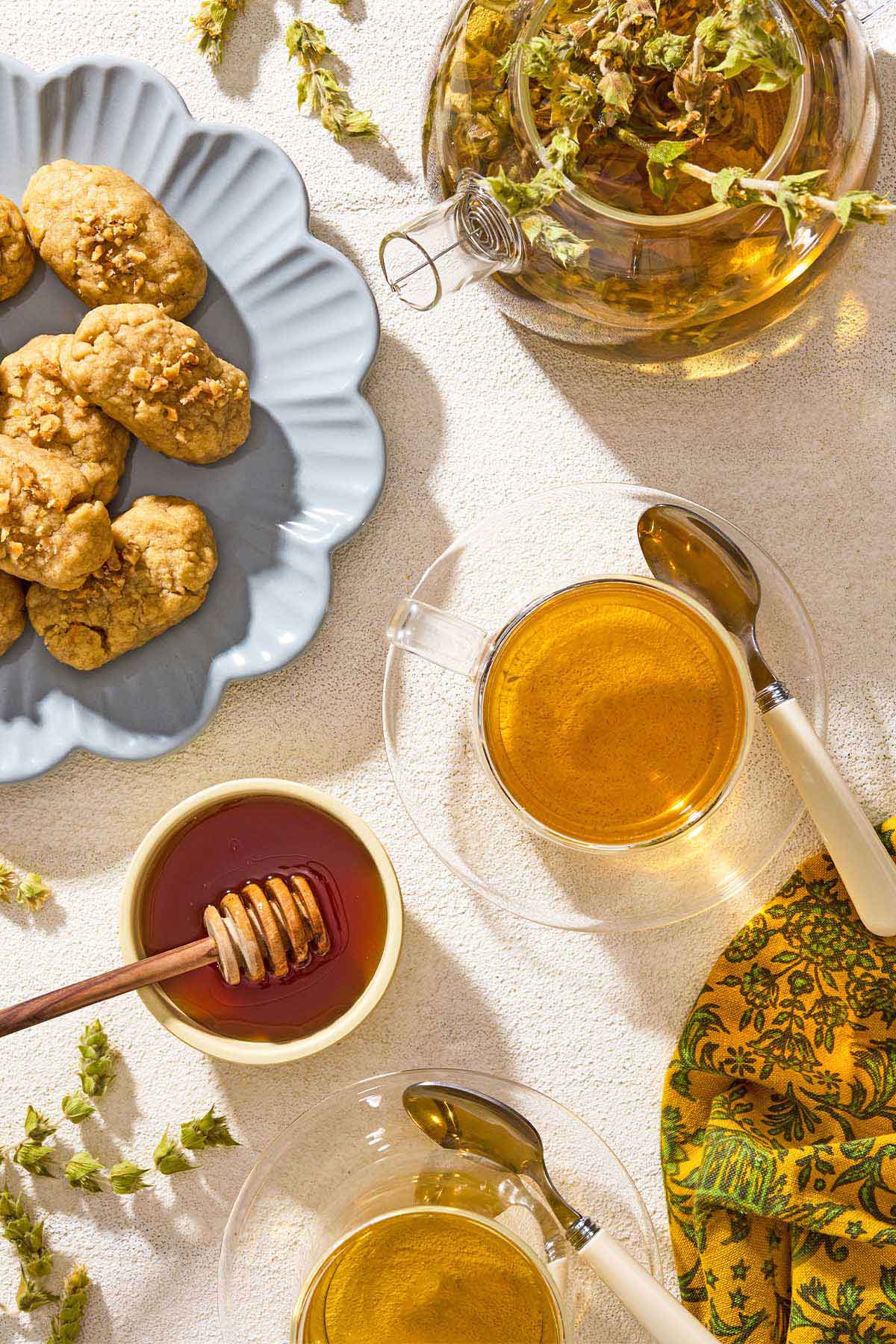 An overhead photo of 2 cups of Greek mountain tea in cups on saucers with spoons. Next to these is a bowl of honey, a kettle of the tea and a plate of cookies.