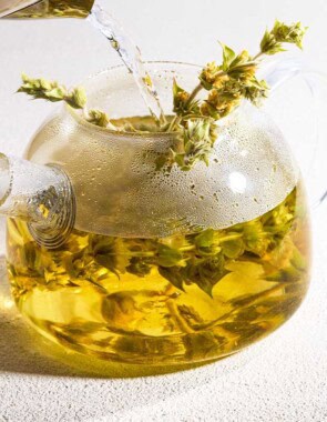 A close up of hot water being poured over a bunch of Greek mountain tea resting in a clear glass kettle. Next to this is a spring of the tea bunch.