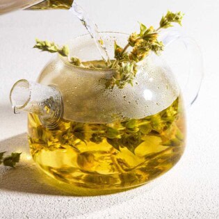 A close up of hot water being poured over a bunch of Greek mountain tea resting in a clear glass kettle. Next to this is a spring of the tea bunch.