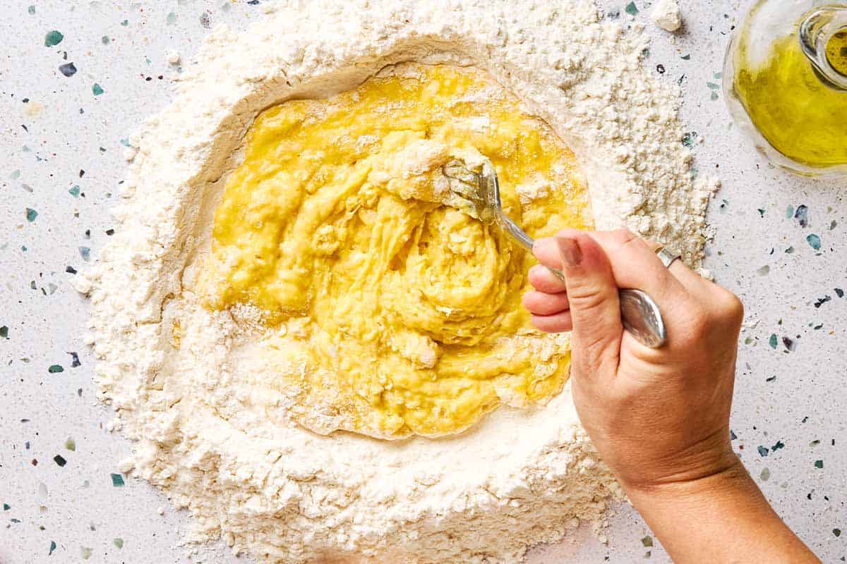 Eggs and olive oil being mixed together with the mound of flour. Next to this is a small jug of olive oil.