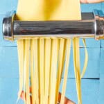 A close up noodles being cut from sheet of homemade pasta dough that's being run through a pasta machine.