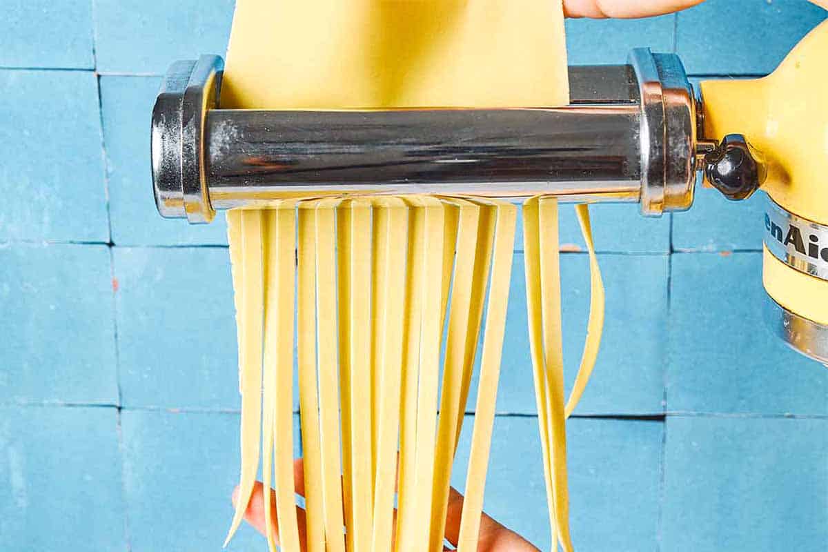 A close up noodles being cut from sheet of homemade pasta dough that's being run through a pasta machine.