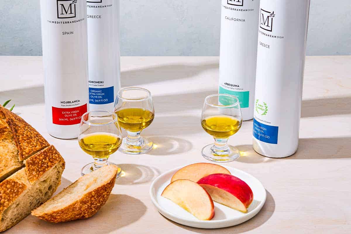 A close up of 4 bottles of various olive oils from The Mediterranean Dish behind 3 glasses of olive oil, a loaf of crusty bread and apple slices on a plate.