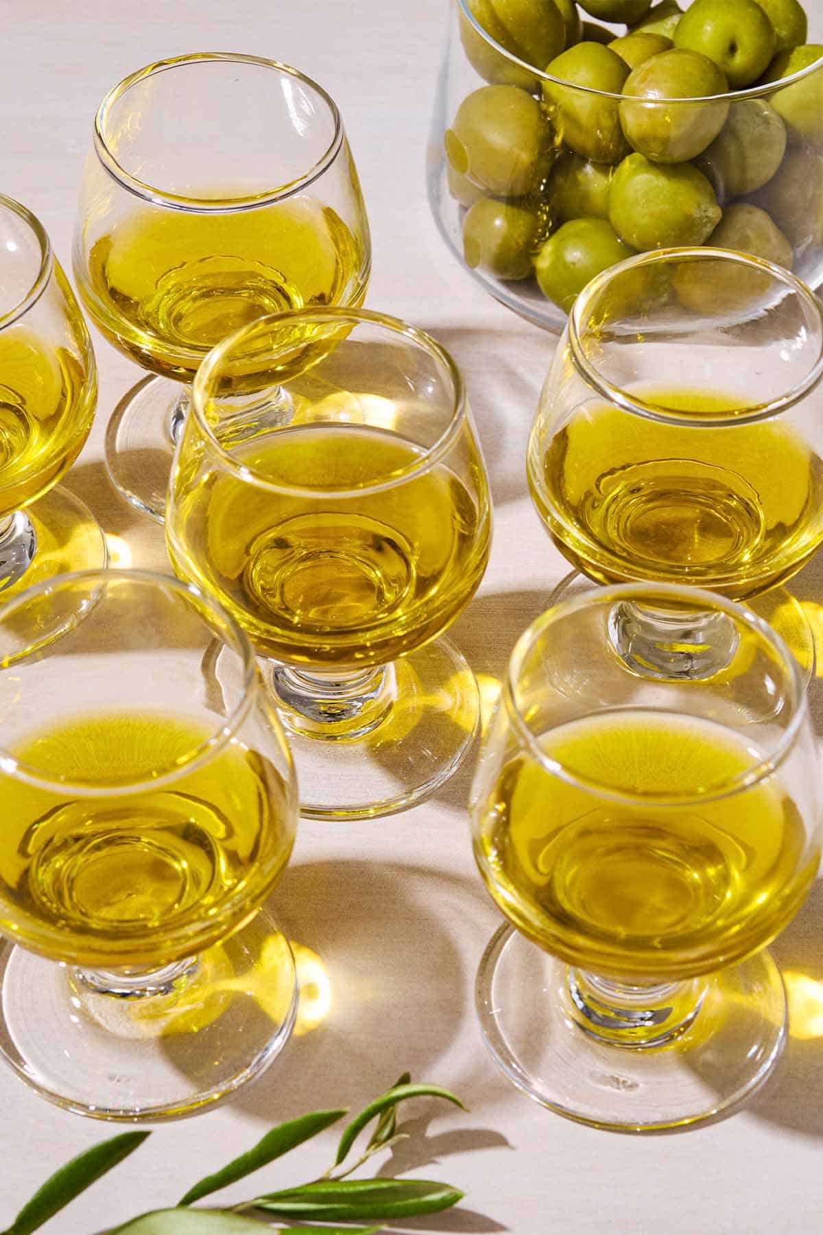 A close up of 6 glasses of various olive oils and a bowl of green olives.